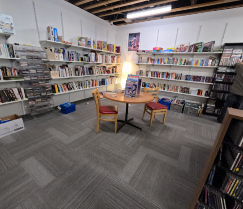 A cozy library space with bookshelves, a round table, and two chairs, illuminated by a warm light.