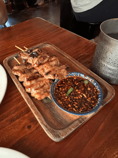 A wooden platter with skewers of grilled meat and a small bowl of spicy dipping sauce.