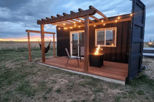 A cozy black container cabin with a wooden deck, chairs, and a fire pit, set against a sunset sky.