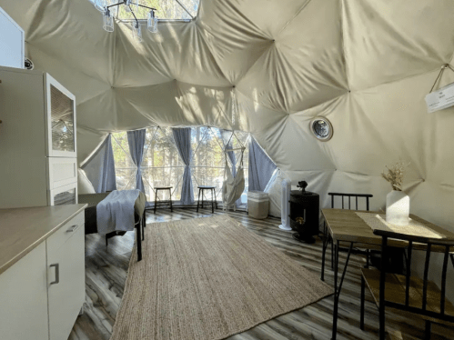 Interior of a geodesic dome with a kitchen area, dining table, and large windows showcasing a natural setting outside.