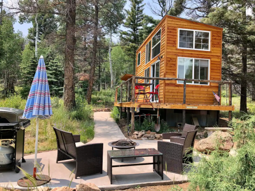A cozy wooden cabin surrounded by trees, featuring a deck, outdoor seating, and a grill in a serene natural setting.