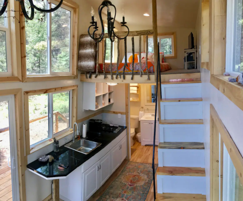 Cozy tiny home interior with a lofted bed, kitchen area, and wooden stairs, surrounded by large windows.