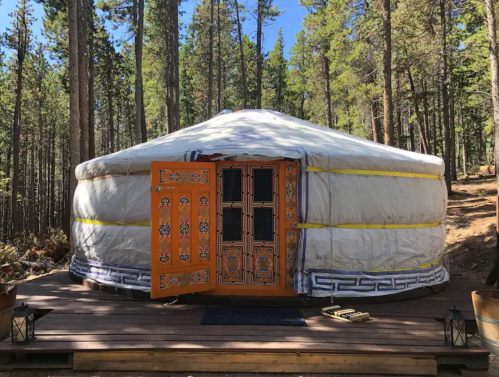 A traditional yurt with a decorative wooden door, set in a forested area with tall trees and a wooden deck.