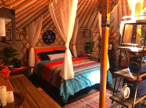 Cozy yurt interior featuring a colorful bedspread, plants, and decorative elements, with warm lighting and wooden accents.