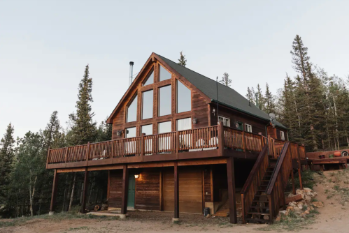 A rustic wooden cabin with large windows, surrounded by tall trees and a deck, set against a clear sky.
