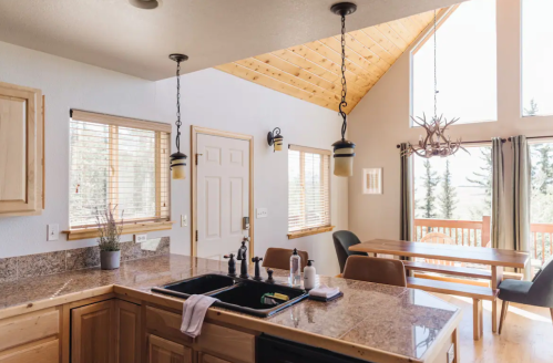 A cozy kitchen with wooden cabinets, a sink, and a dining area featuring large windows and a view of trees outside.