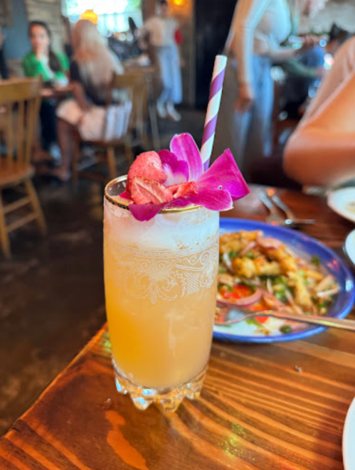 A refreshing cocktail garnished with a pink flower and a straw, placed on a wooden table with food in the background.