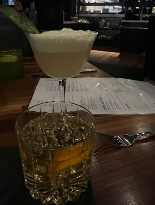 A cocktail glass with a frothy drink on top and a whiskey glass with an orange slice, set on a wooden table.