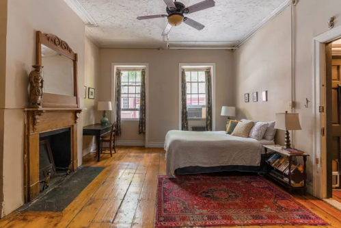 Cozy bedroom with a bed, wooden floors, a fireplace, and large windows, featuring a rug and a small desk.