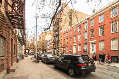 A city street lined with brick buildings and parked cars, featuring trees and pedestrians in the background.