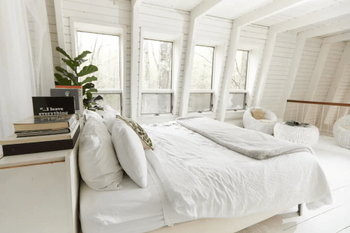Cozy bedroom with a white bed, large windows, and a plant, featuring a minimalist design and natural light.