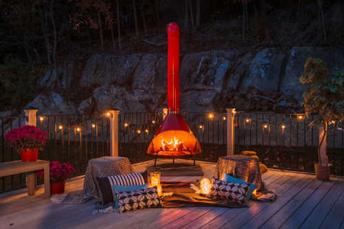 Cozy outdoor space with a red fireplace, blankets, candles, and flowers, illuminated by string lights at dusk.