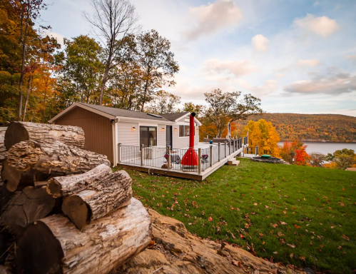 A cozy cabin with a deck overlooking a lake, surrounded by autumn foliage and stacked logs in the foreground.