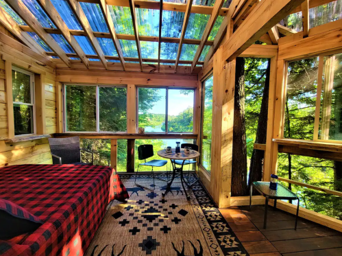 Cozy cabin interior with a bed, table, and large windows overlooking a lush forest and lake.