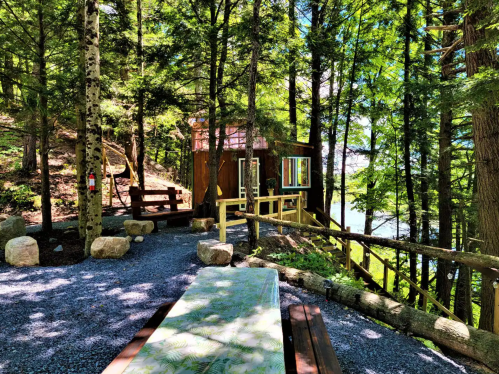 A cozy cabin nestled among trees, with a gravel path, wooden benches, and a view of a lake in the background.