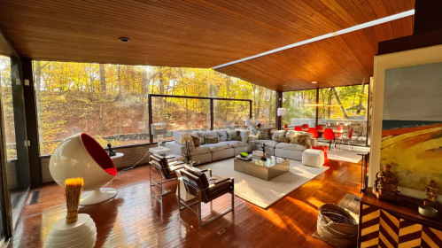 Modern living room with large windows, wooden ceiling, stylish furniture, and autumn scenery outside.