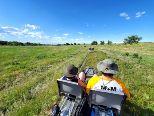 Two people in hats ride on a rail cart through a grassy field under a blue sky with scattered clouds.
