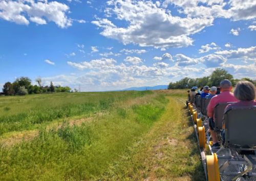 A group of people on a train-like vehicle travels along a grassy path under a blue sky with fluffy clouds.