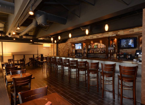 A modern bar area with wooden stools, a well-stocked bar, and two TVs in a stylish restaurant setting.