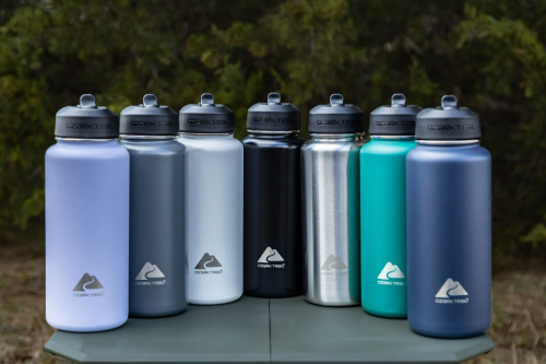 A row of eight colorful water bottles on a table, featuring various designs and sizes, set against a natural background.