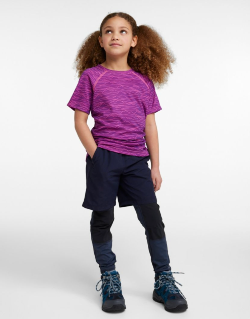 A young girl with curly hair stands confidently in a purple athletic shirt and navy shorts, wearing hiking boots.
