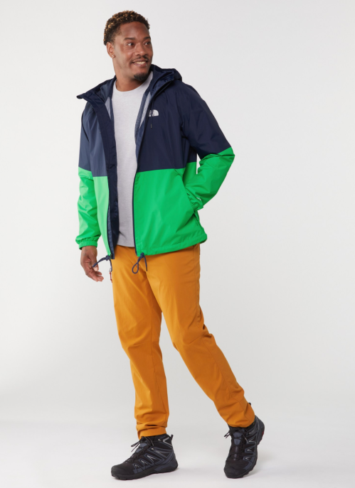 A man in a colorful jacket and orange pants stands smiling against a plain background.