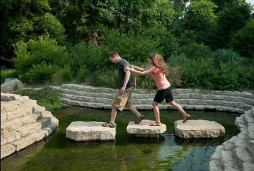 A man carefully steps on stones across a pond while a woman playfully pulls him forward in a lush garden setting.