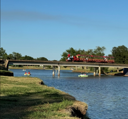 A red train crosses a bridge over a river, with two kayakers paddling nearby under a clear blue sky.