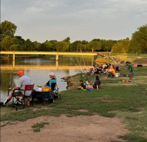 A group of people fishing along a riverbank, with trees and a bridge in the background under a partly cloudy sky.