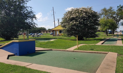 A mini-golf course with green turf and blue obstacles, surrounded by trees and a building in the background.