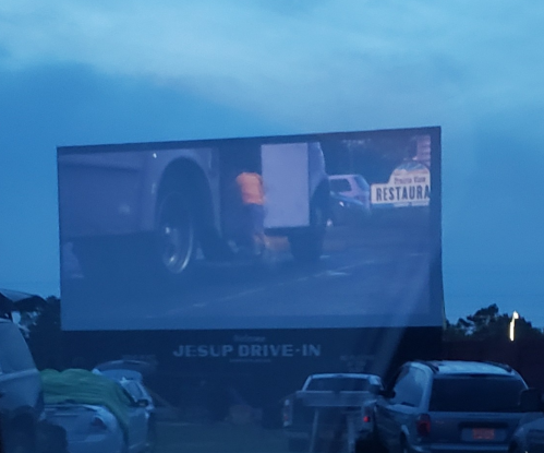 A drive-in movie screen shows a scene with a person near a vehicle, surrounded by parked cars at dusk.