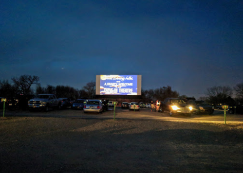A drive-in theater at dusk, with cars parked and a large screen displaying a welcome message.