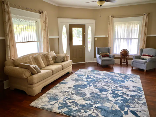 Cozy living room with a beige sofa, two armchairs, and a floral rug, featuring large windows and a front door.