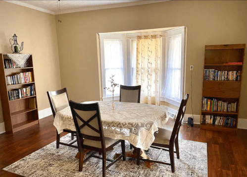 A cozy dining area with a round table, chairs, a lace tablecloth, and bookshelves filled with books. Natural light filters in.