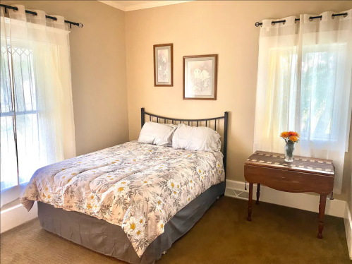 Cozy bedroom with a double bed, floral bedding, wooden side table, and natural light from two windows.