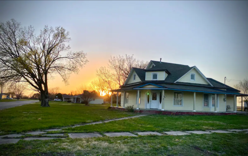 A charming yellow house at sunset, surrounded by green grass and trees, with a clear sky in the background.