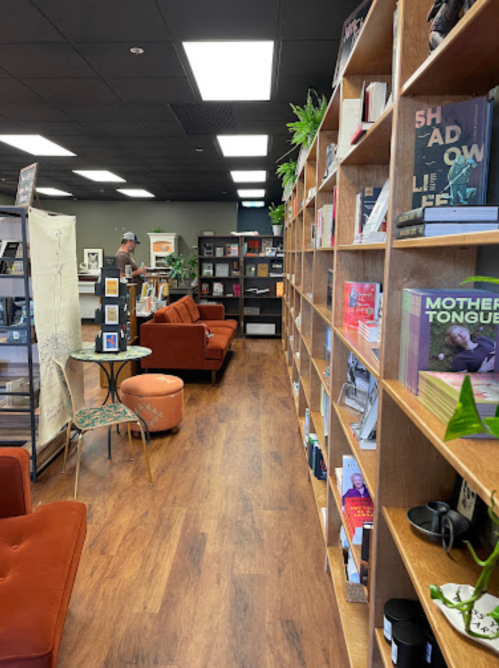 A cozy bookstore interior with wooden shelves filled with books, a reading area, and a person browsing in the background.