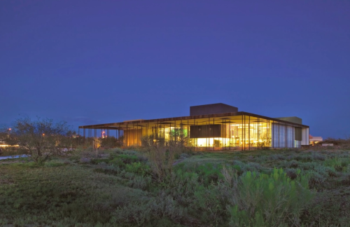 Modern building with large glass windows, surrounded by greenery, illuminated at dusk against a twilight sky.