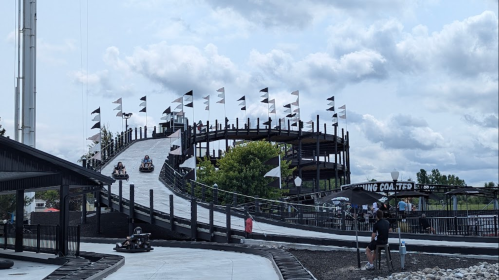 A go-kart track with a steep incline, featuring black and white flags, surrounded by trees and cloudy skies.