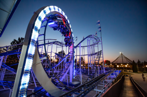 A roller coaster with bright lights loops against a twilight sky, with a tent structure visible in the background.