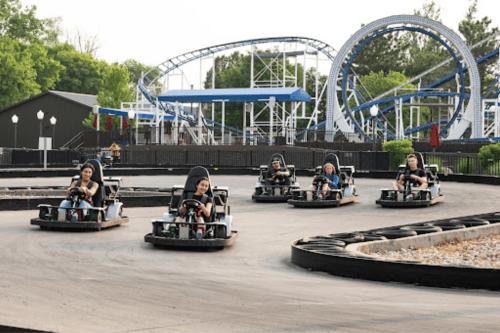 A group of children racing go-karts on a track with a roller coaster in the background.