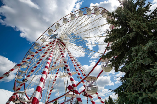 A colorful ferris wheel with red and white stripes against a blue sky with fluffy clouds and a green tree nearby.