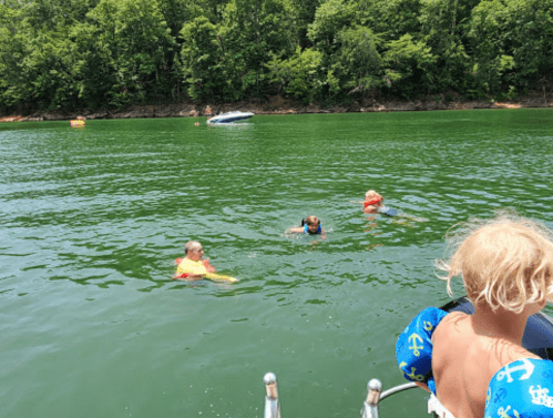 Three people swimming in a green lake, with a boat in the background and a child in a life jacket nearby.
