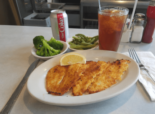 A plate of fried fish fillets with a lemon wedge, served with broccoli, green beans, and a drink on the side.