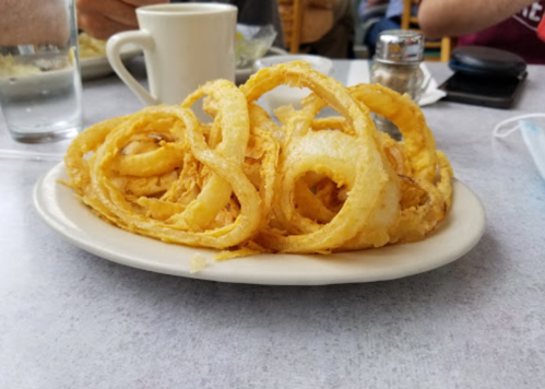 A plate of golden, crispy onion rings served on a white dish, with a cup and other table items in the background.