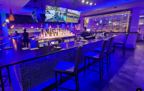 A modern bar with blue lighting, high stools, and two large screens displaying images. Staff member is behind the bar.
