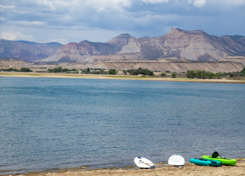 A serene lake with sandy shore, paddleboards, and kayaks, surrounded by mountains under a cloudy sky.