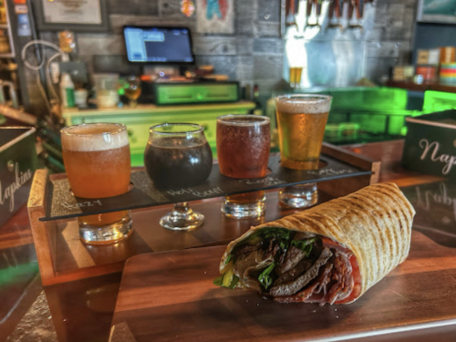 A wrap filled with meat and greens, with a flight of four craft beers in the background on a wooden tray.