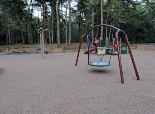 A playground with a swing set, slide, and climbing structure surrounded by trees and a gravel path.