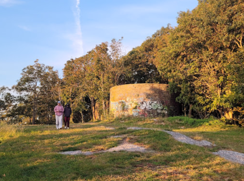 Two people stand near a stone structure surrounded by trees, with graffiti visible on the structure, under a clear sky.